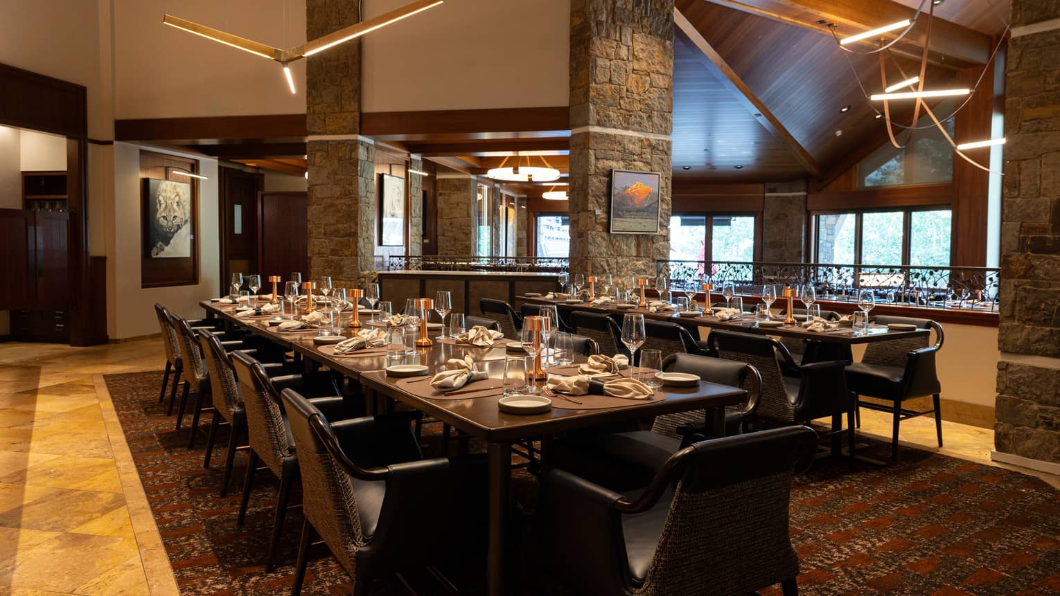 A warm interior dining space with windows on one wall, wood floors, long dining tables set for a meal, chairs, and large three-bladed ceiling fans suspended from the high ceiling, Four Seasons Resort Jackson Hole, Teton Village, USA