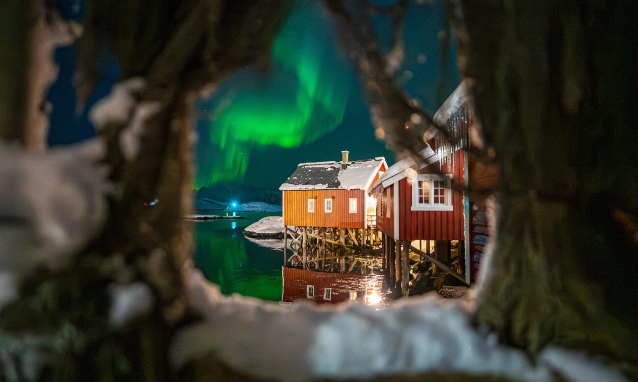 Red waterfront cabin beneath green northern lights, framed by dark rocks at Svinøya Rorbuer, Lofoten, Norway