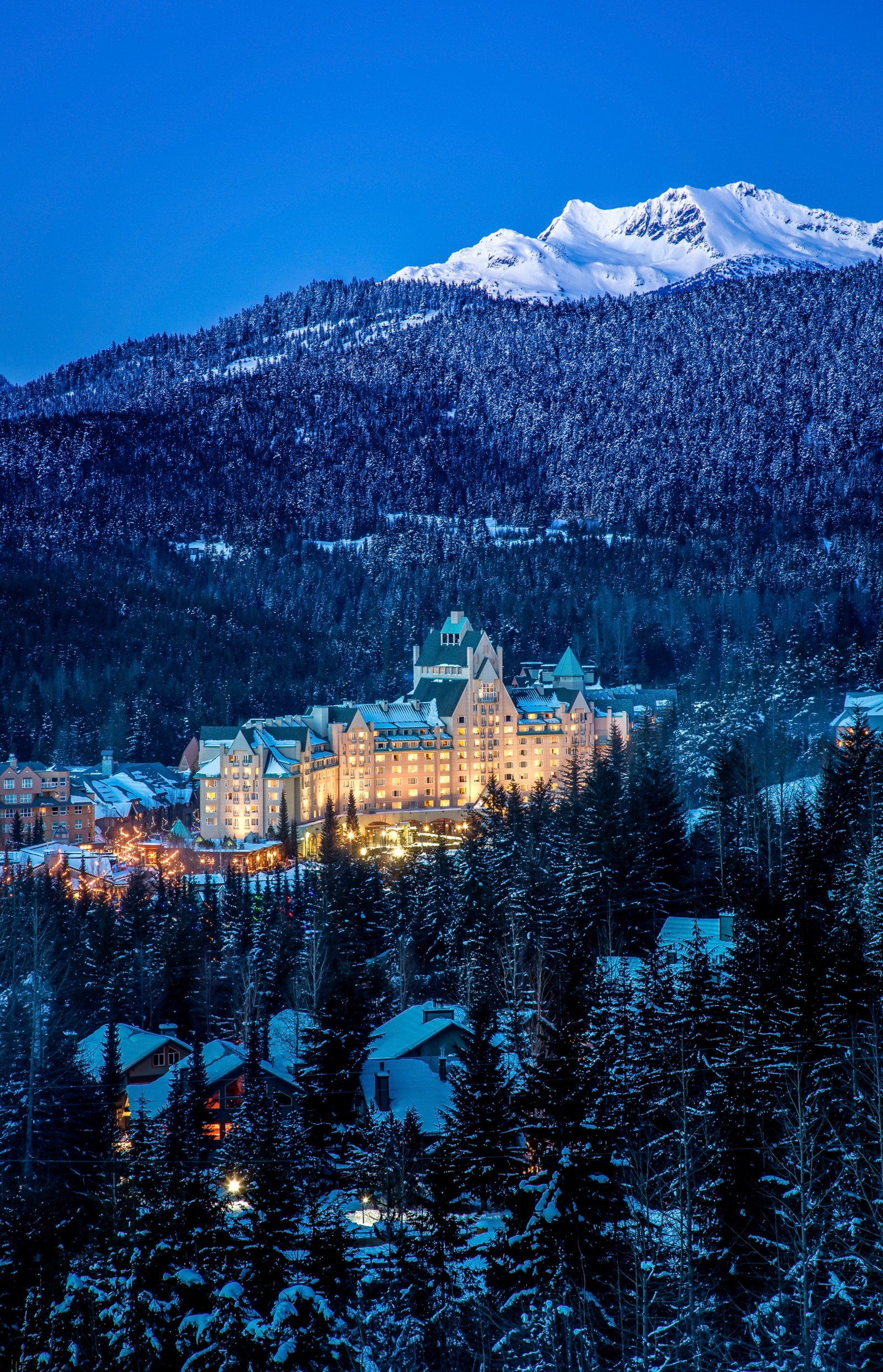 Illuminated Fairmont Chateau Whistler amid snow-covered forest beneath mountain peaks at blue hour, British Columbia, Canada