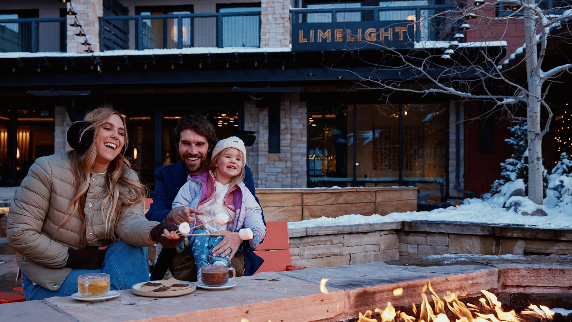 Family toasting marshmallows beside an outdoor fire pit at Limelight Hotel Snowmass in winter.