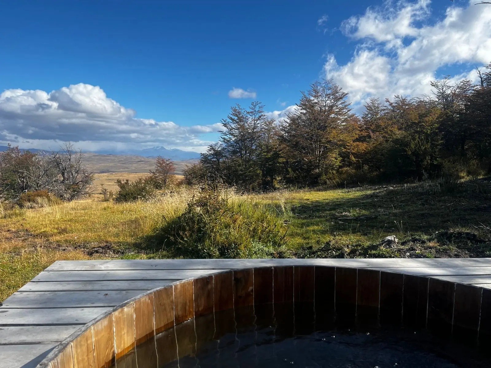 Round wooden outdoor hot tub beside a villa deck, overlooking grassland, trees and distant mountains, Awasi Patagonia