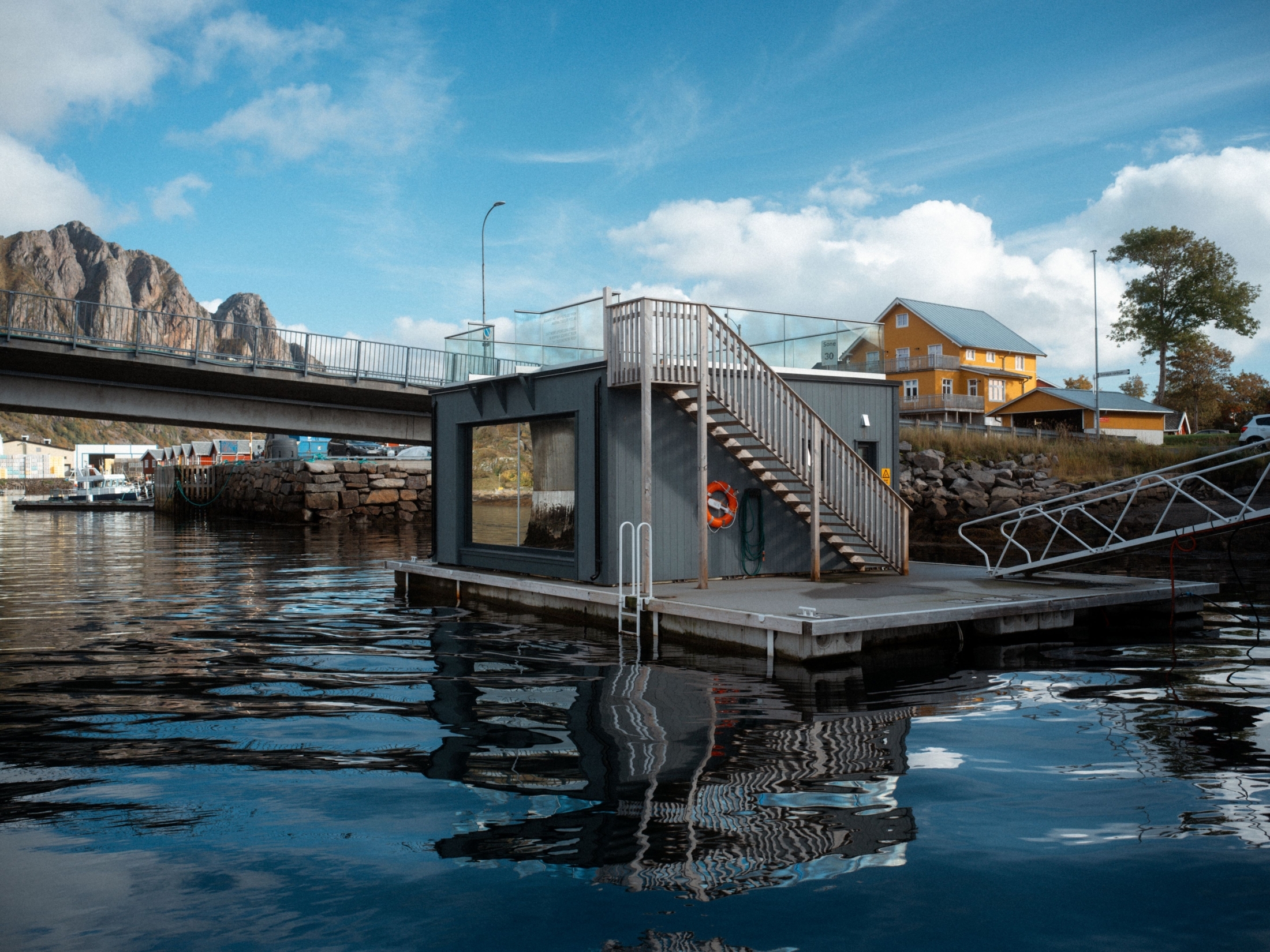 Floating sauna moored beside a bridge with an outdoor staircase and harbour reflections.
