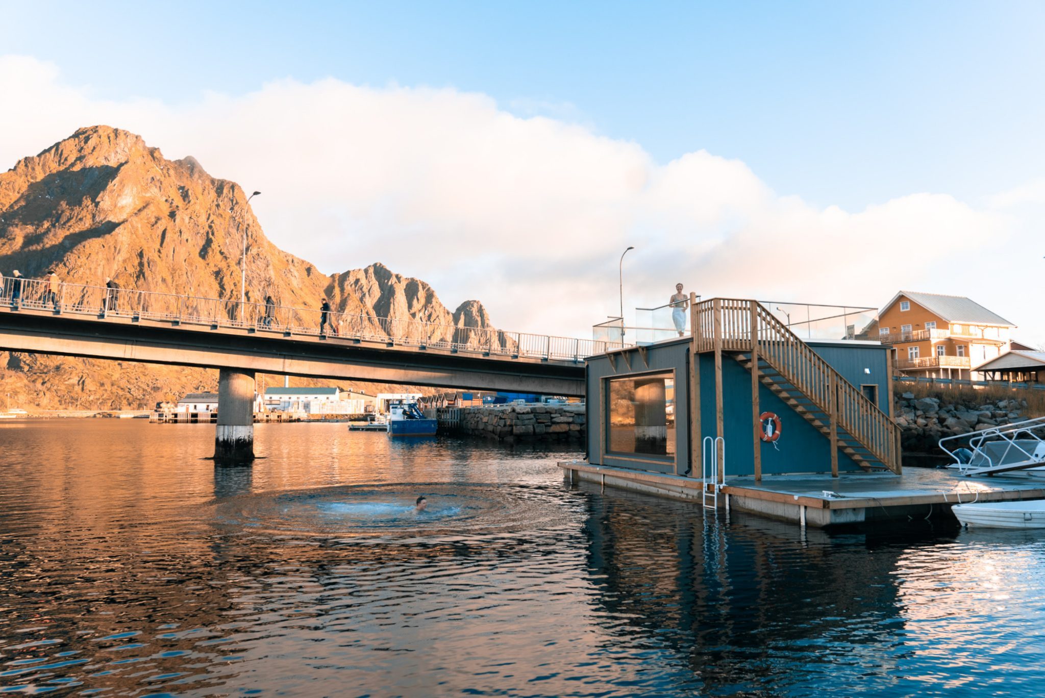 Floating sauna and harbour bridge in warm light beneath steep mountains.