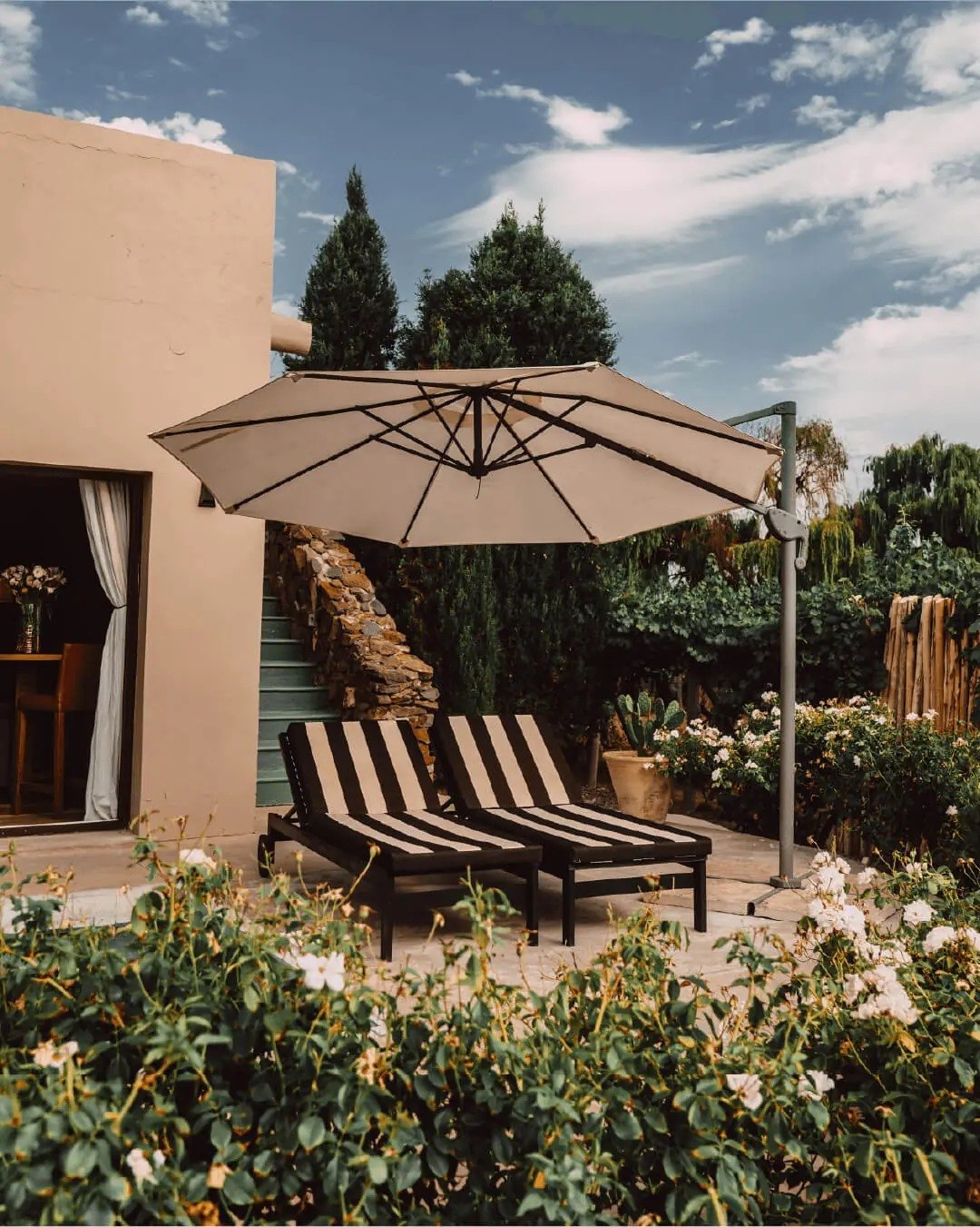 Striped loungers beneath a patio umbrella beside a pale villa wall and flowering garden, Cavas Wine Lodge