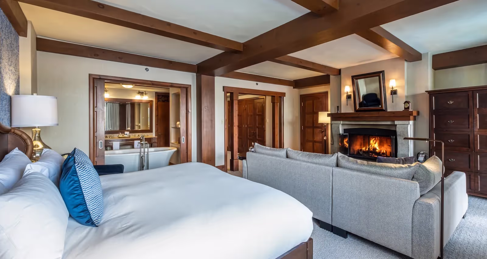 Suite with a king bed, pale upholstered seating, exposed ceiling beams and a lit stone fireplace at Hôtel Quintessence