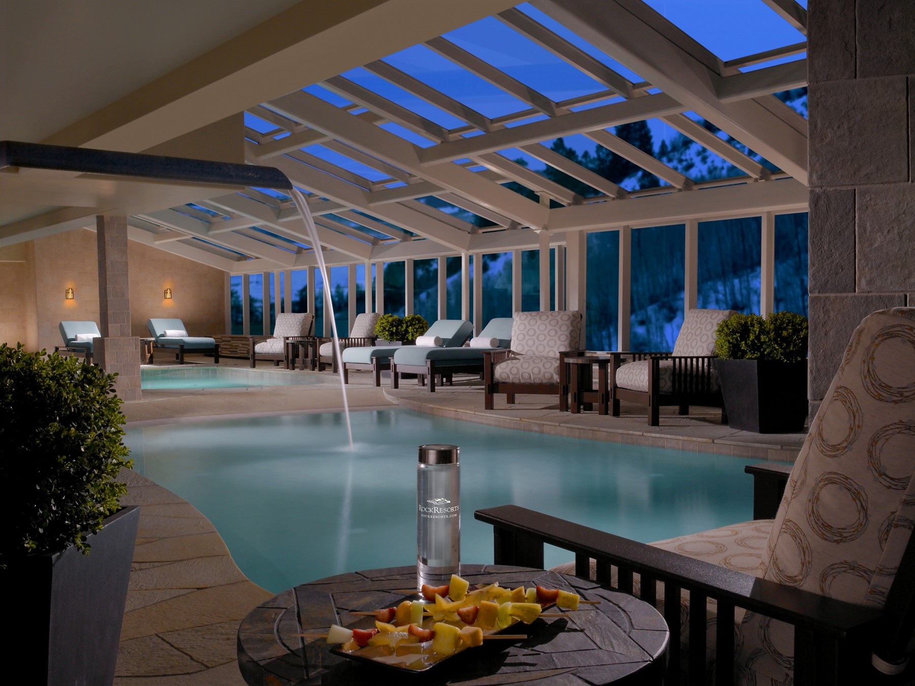Indoor swimming pool at One Ski Hill Place beneath skylights beside large windows at dusk.