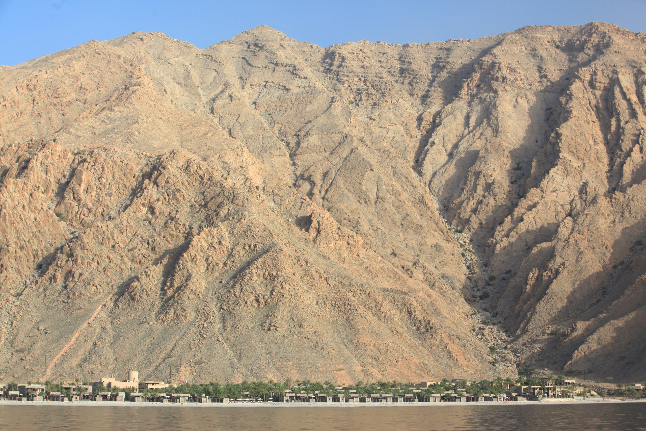 Six Senses Zighy Bay village and palms at the foot of a steep mountain, viewed from the sea.