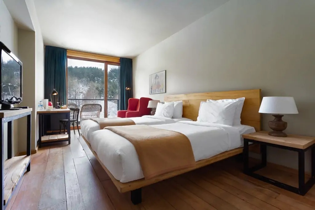 Twin beds on a wooden platform with a red armchair and forest-facing window at Rooms Hotel Kazbegi, Georgia