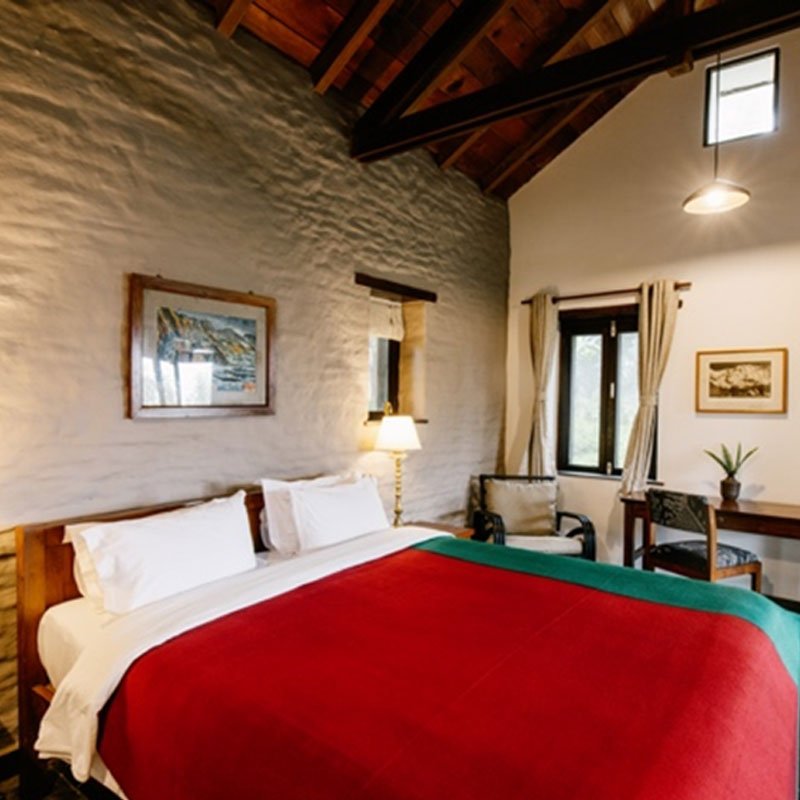 Cottage bedroom with white walls, exposed rafters and a bed dressed with a red cover at Tiger Mountain Pokhara Lodge, Nepal