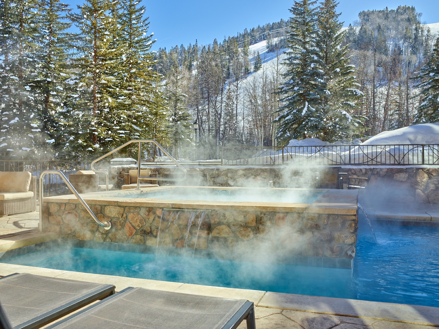 Steaming outdoor pool and hot tub beneath snowy trees at The Osprey at Beaver Creek.