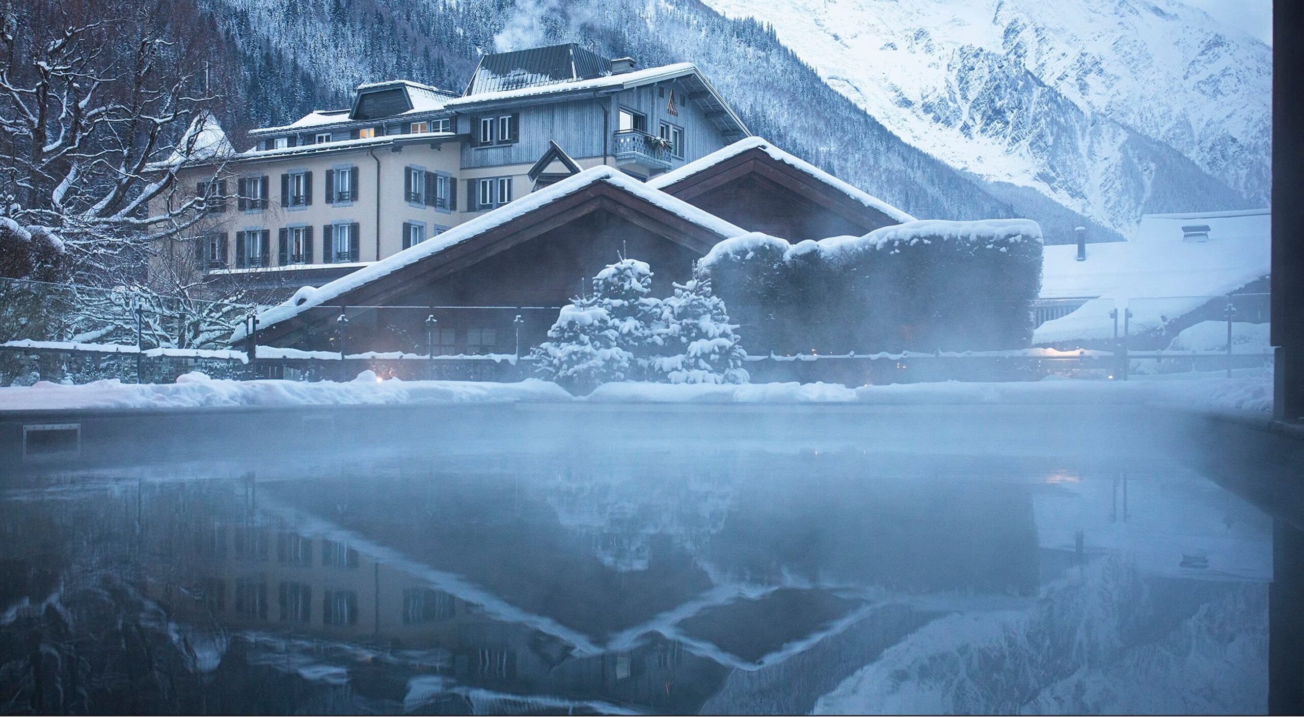 Steam rising from the outdoor swimming pool beneath snow-covered chalets and Alpine peaks, Le Hameau Albert 1er, Chamonix, France