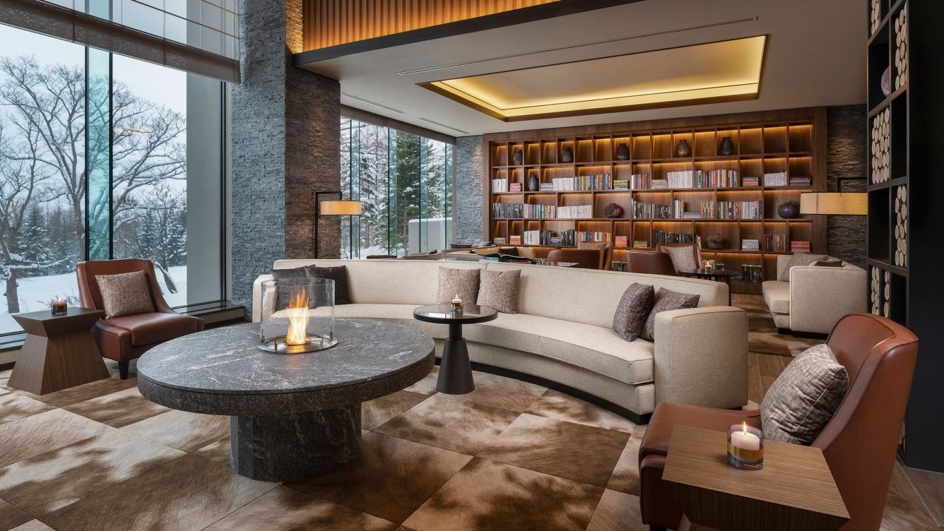 Ume Lounge with upholstered seating, a round table, bookshelves and large windows facing leafless trees, Higashiyama Niseko Village, a Ritz-Carlton Reserve