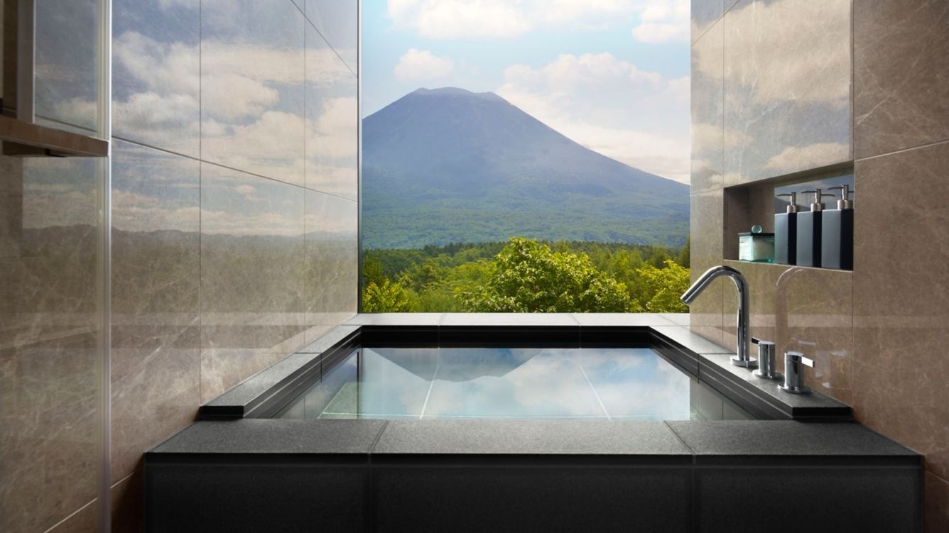Rectangular soaking tub beside a panoramic bathroom window overlooking green trees and Mount Yotei, Higashiyama Niseko Village, a Ritz-Carlton Reserve