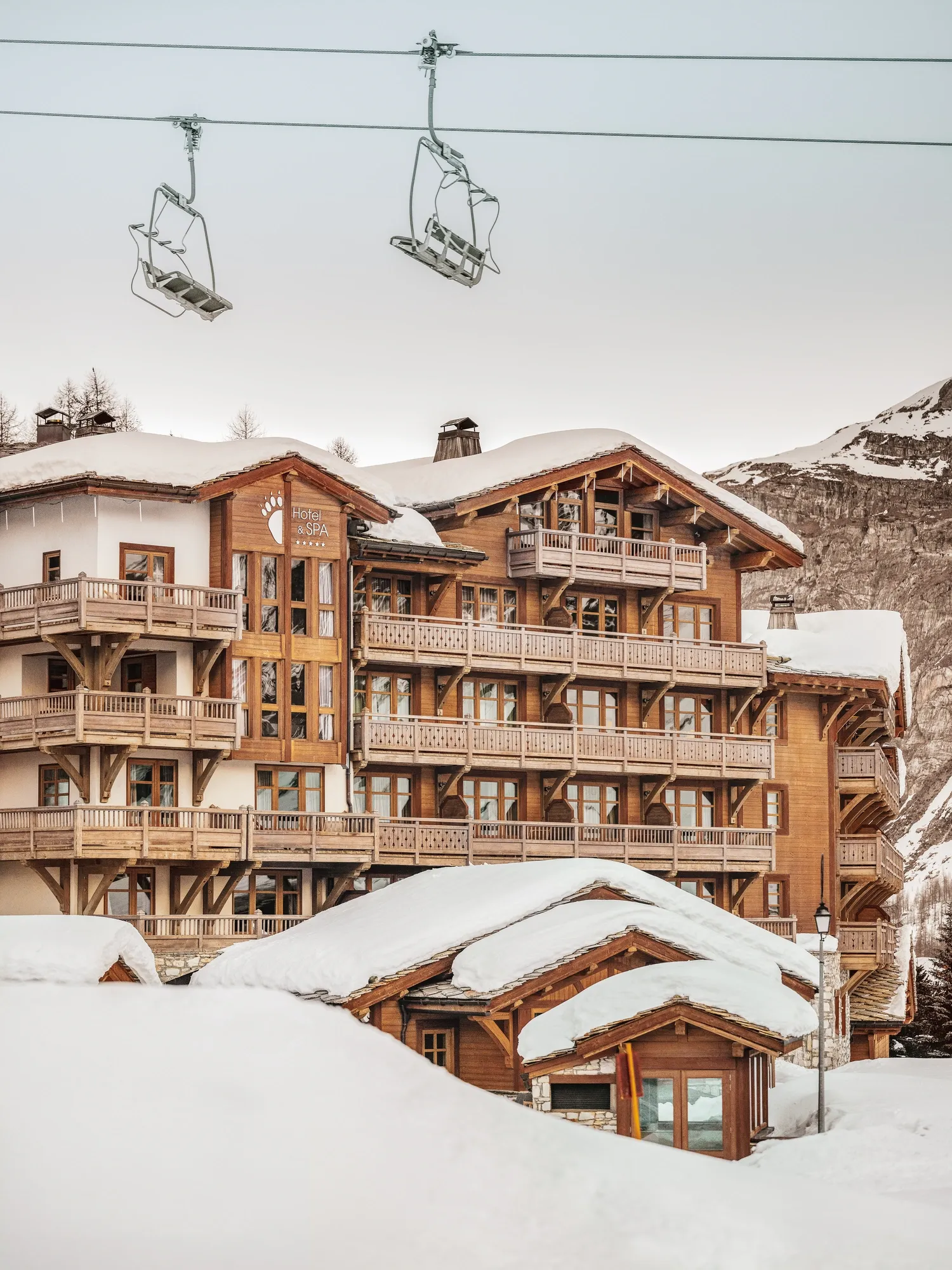 Snow-covered timber-and-stone exterior of Les Barmes de l’Ours beneath the ski lifts and slopes of Bellevarde, Val d’Isère, France