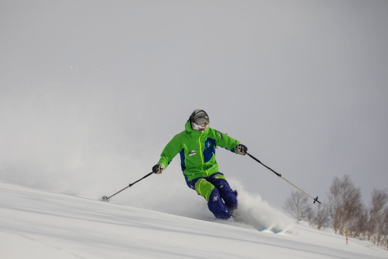 Skier in a bright green jacket making a turn on a snowy slope, throwing up powder beneath an overcast sky, Higashiyama Niseko Village, a Ritz-Carlton Reserve