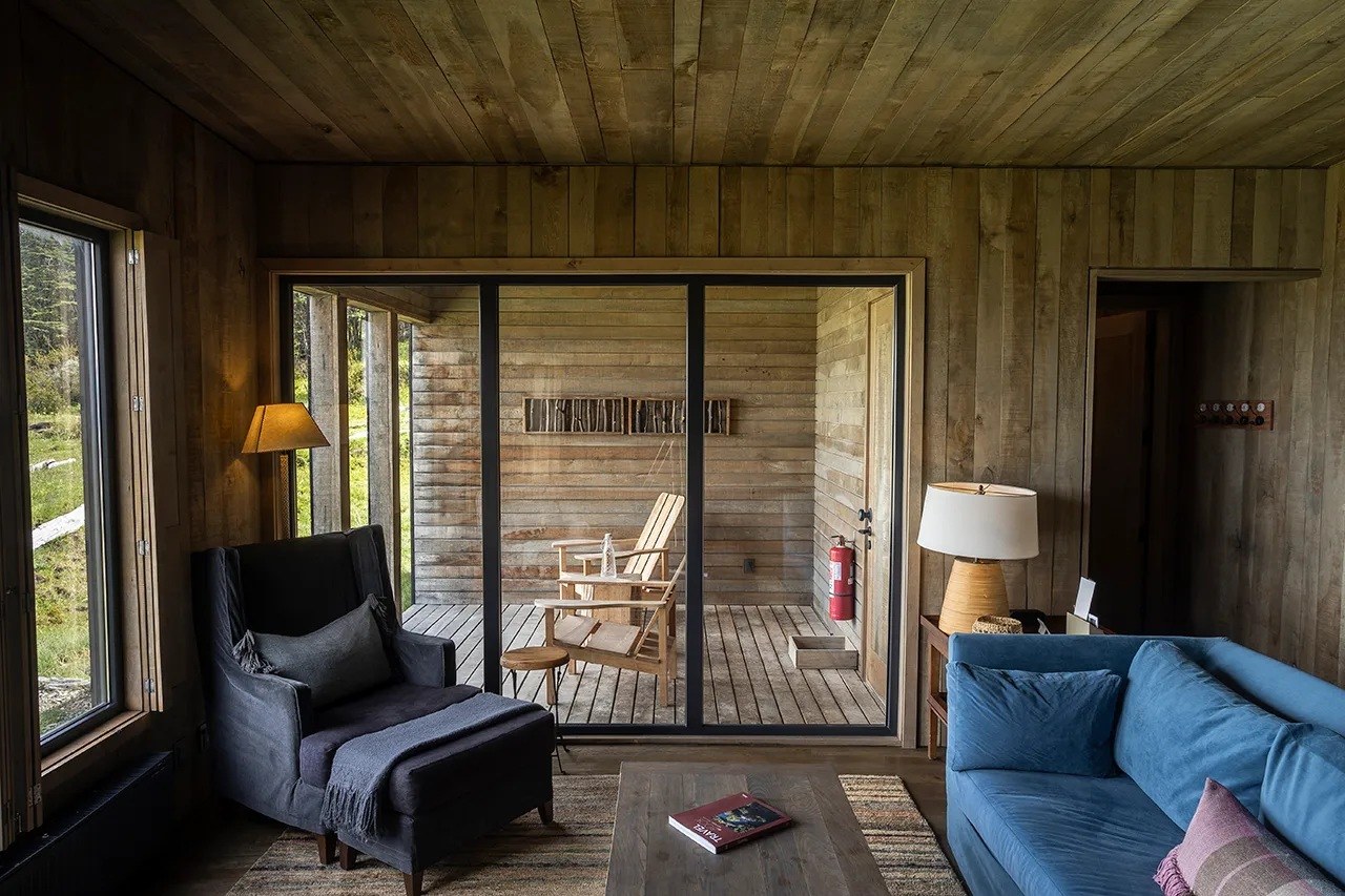Wood-paneled villa sitting area with a blue sofa, armchair and glass doors to a terrace, Awasi Patagonia