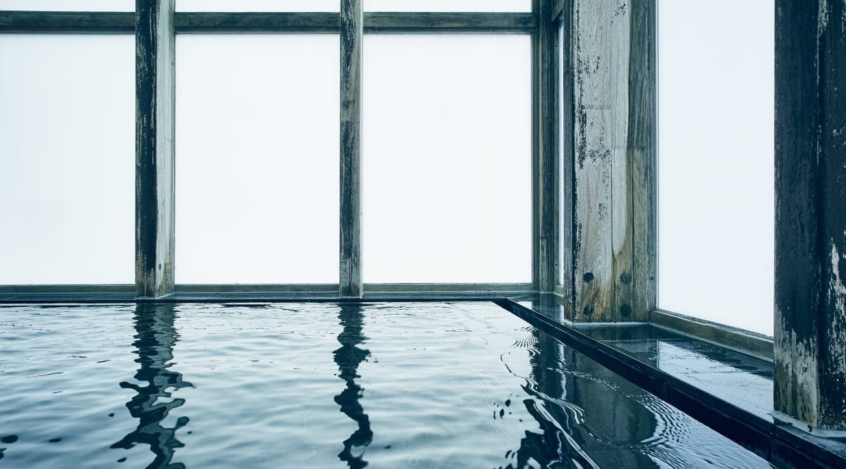 Meditation Bath interior: rippling hot-spring water beside tall frosted windows and weathered dark timber posts, Hoshinoya Karuizawa
