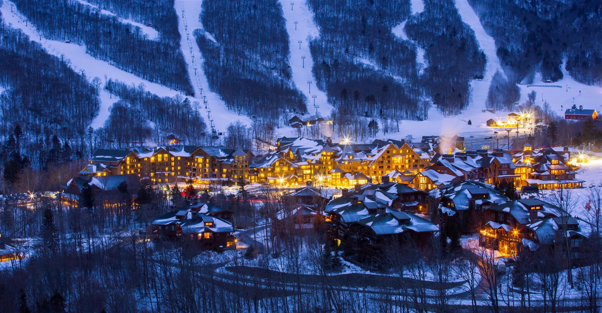 Snowy resort village and illuminated lodge beneath ski slopes, The Lodge at Spruce Peak, Stowe, USA