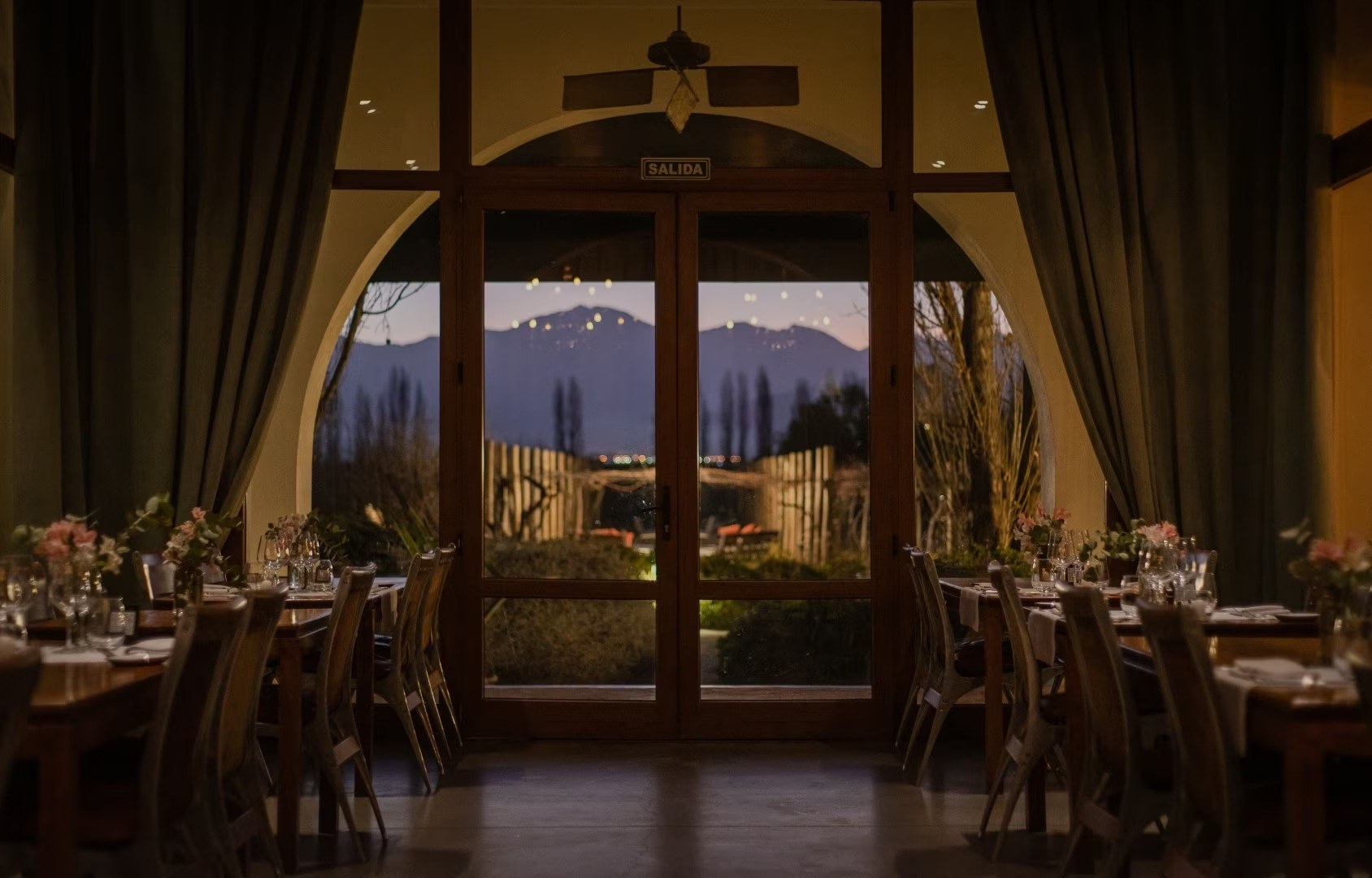 Restaurant tables beside tall arched windows framing vineyards and distant mountains at dusk, Cavas Wine Lodge