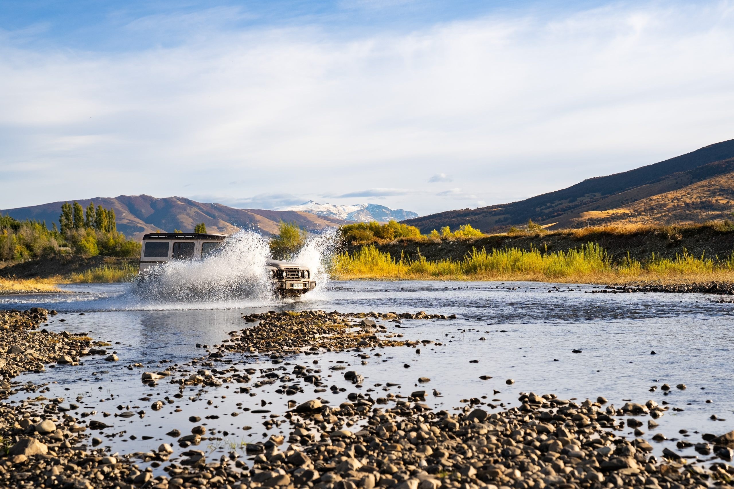 Four-wheel-drive vehicle crossing a shallow river with a spray of water against a mountain backdrop, Awasi Patagonia