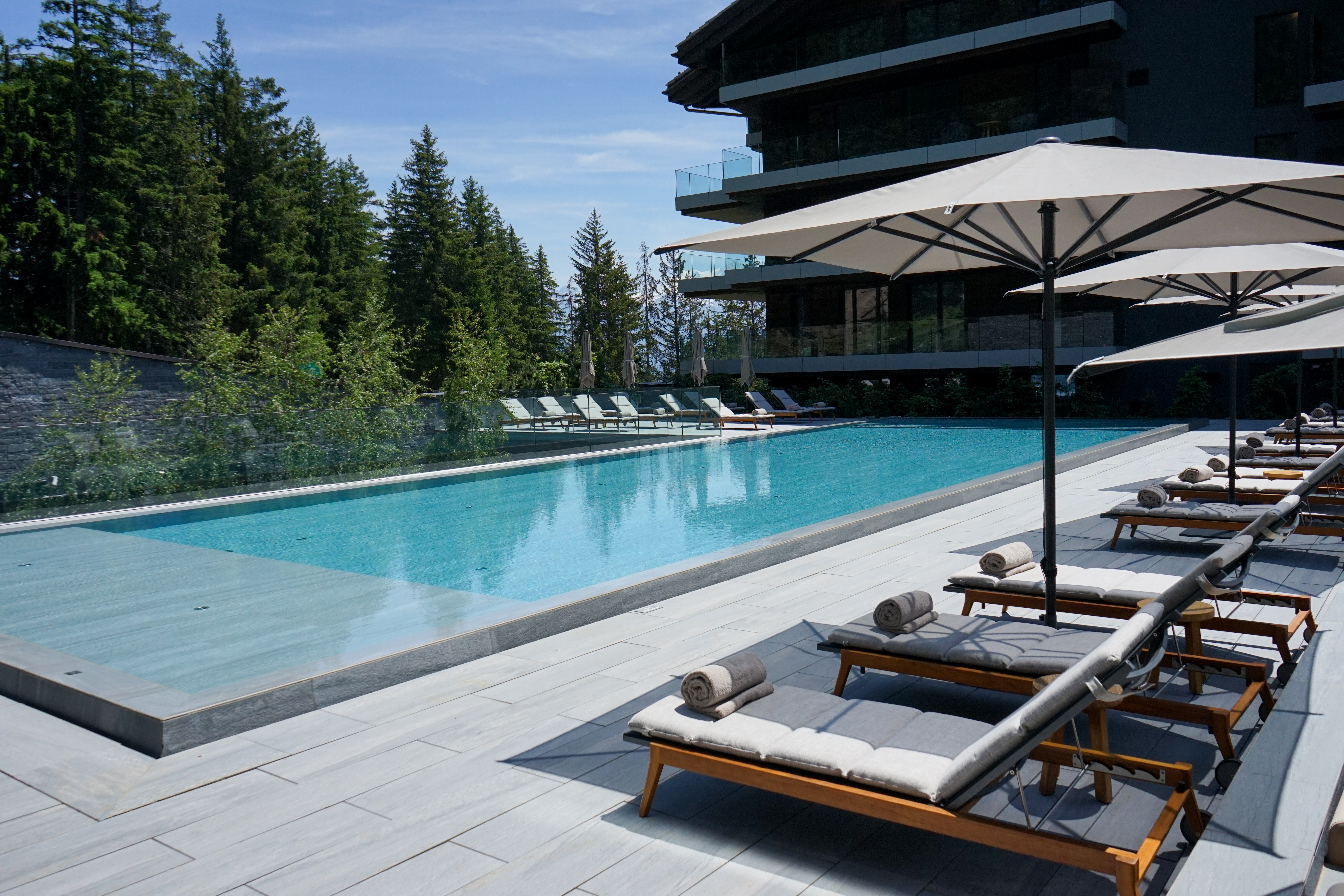 Hotel exterior and outdoor pool with loungers beside Alpine forest in summer, Six Senses Crans-Montana, Crans-Montana, Switzerland